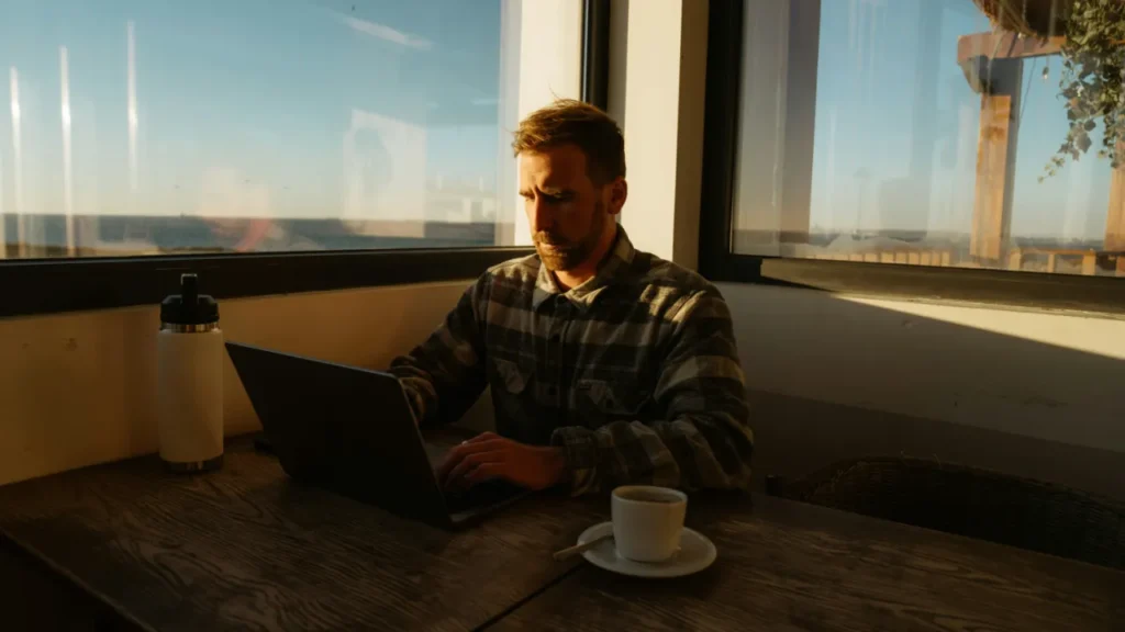 12:22 AM A man sits at a cafe table with a laptop and coffee, studying English — used in a B1 lesson plan on thinking in English.