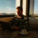 12:22 AM A man sits at a cafe table with a laptop and coffee, studying English — used in a B1 lesson plan on thinking in English.