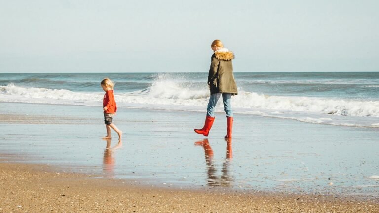 A woman in red boots walks along the beach beside a child playing near the water. Image for an A2 ESL lesson from betterclass comparing childhood and adulthood, focusing on memories, responsibilities, and daily life.