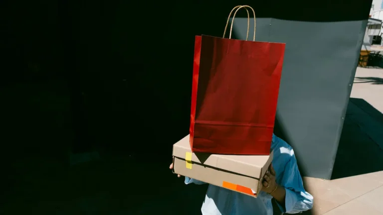 A person holding a large red shopping bag and a box in front of their face while standing outdoors; image used for an A2 ESL lesson plan about asking for a discount.