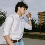 A young man in a white shirt and jeans walks through a park with a tote bag and coffee cup — a visual from the Performative Male B2 ESL lesson plan exploring masculinity, social media image, and authenticity in modern English learning.