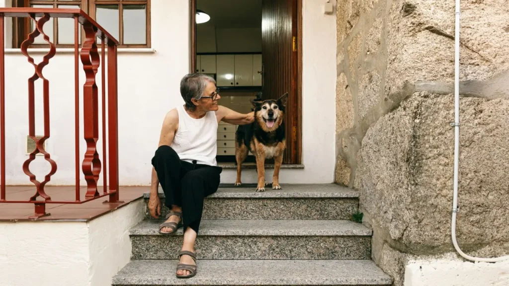 An elderly woman sits on her front steps smiling beside her dog. The image represents the What Does Aging Really Feel Like? B2 ESL lesson from betterclass, where students explore how aging affects the body and mind through vocabulary, video analysis, and reflective discussions about empathy, independence, and life experience.