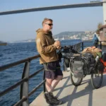 Man with sunglasses stands by a loaded touring bike beside the water, with a large bridge and ferry visible in the background.