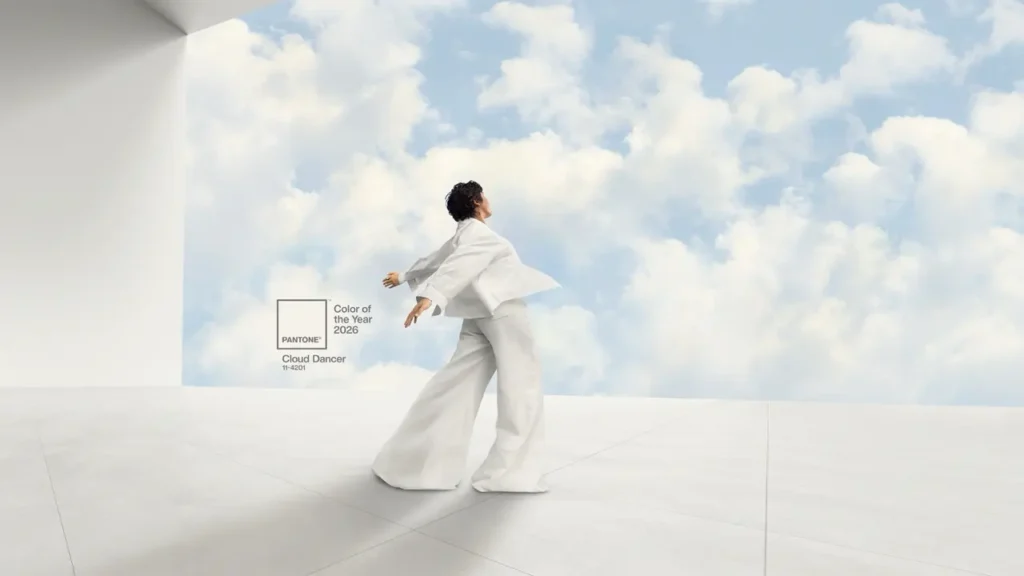 A person wearing a flowing white outfit stands against a sky background with billowing clouds, with a Pantone label showing 'Color of the Year 2026 Cloud Dancer 11-4201' displayed in the image.