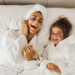 Mother and young daughter wearing white bathrobes and purple face masks, smiling together while doing skincare routine