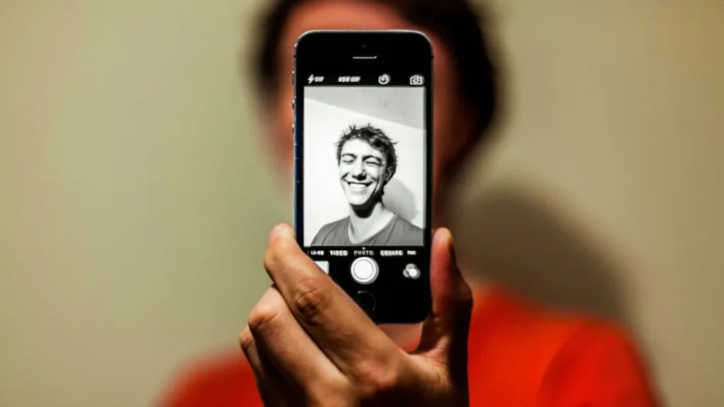 Person holding a smartphone showing a black-and-white selfie on the screen, with the person blurred in the background.