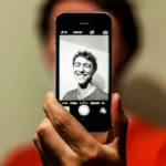 Person holding a smartphone showing a black-and-white selfie on the screen, with the person blurred in the background.