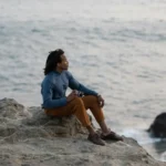 A solitary figure sits on seaside rocks overlooking the ocean, symbolizing modern singlehood and independence