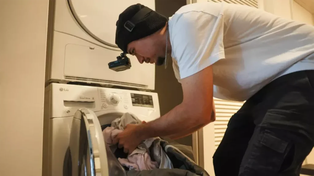 Man wearing a head-mounted camera loads laundry into a washing machine, filming daily chores to train robots for esl news articles lesson.