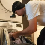 Man wearing a head-mounted camera loads laundry into a washing machine, filming daily chores to train robots for esl news articles lesson.