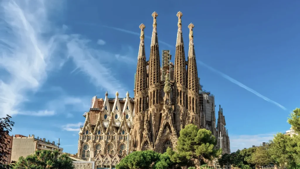 A daytime exterior view of the Sagrada Família in Barcelona, with its tall stone spires rising against a bright blue sky.