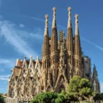 A daytime exterior view of the Sagrada Família in Barcelona, with its tall stone spires rising against a bright blue sky.