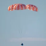 The Orion capsule descends under three red and white parachutes toward the Pacific Ocean during Artemis II splashdown.