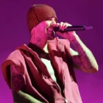 Justin Bieber singing into a microphone on stage, lit by purple light. He is wearing a red beanie hat and a pink zip-up vest, with tattoos visible on his neck and arms.