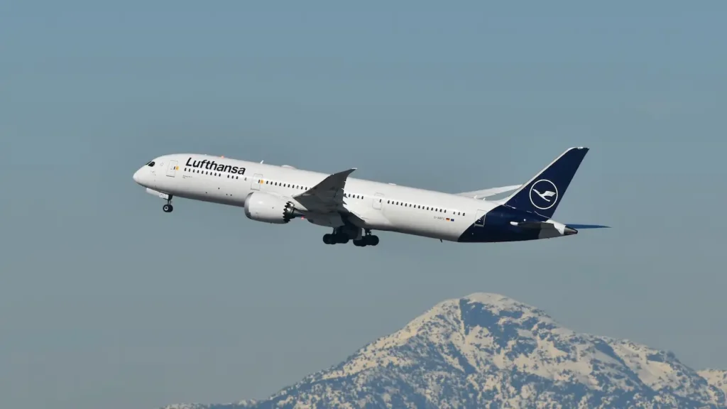A Lufthansa passenger jet climbs away from a mountain backdrop against a clear sky.