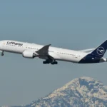 A Lufthansa passenger jet climbs away from a mountain backdrop against a clear sky.