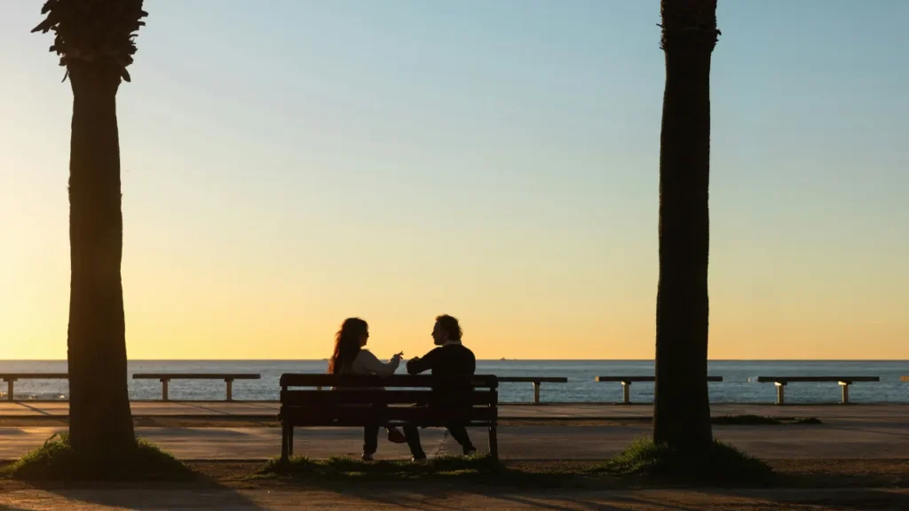 Two people sitting on a bench by the sea at sunset, deep in conversation — B2 lesson plan on asking meaningful questions.