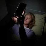 A woman lies in bed in a dark room scrolling on her phone late at night, her face lit by the screen in a phone addiction lesson.