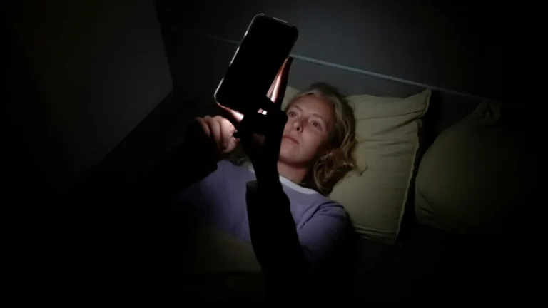A woman lies in bed in a dark room scrolling on her phone late at night, her face lit by the screen in a phone addiction lesson.