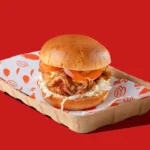 Crispy chicken burger with tomato and slaw in a cardboard tray on a bright red background, styled like a fast food ad.