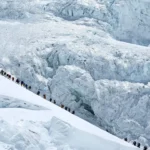 A long line of climbers in colorful gear ascending a steep snow-covered ridge on Mount Everest, with massive glacial ice walls in the background.