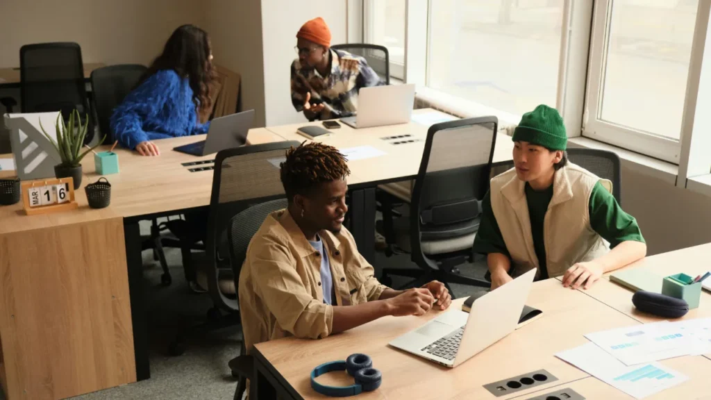 Two colleagues discussing work at a shared office desk -- a B2 lesson plan feedback activity in a modern workplace setting.