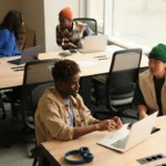 Two colleagues discussing work at a shared office desk -- a B2 lesson plan feedback activity in a modern workplace setting.