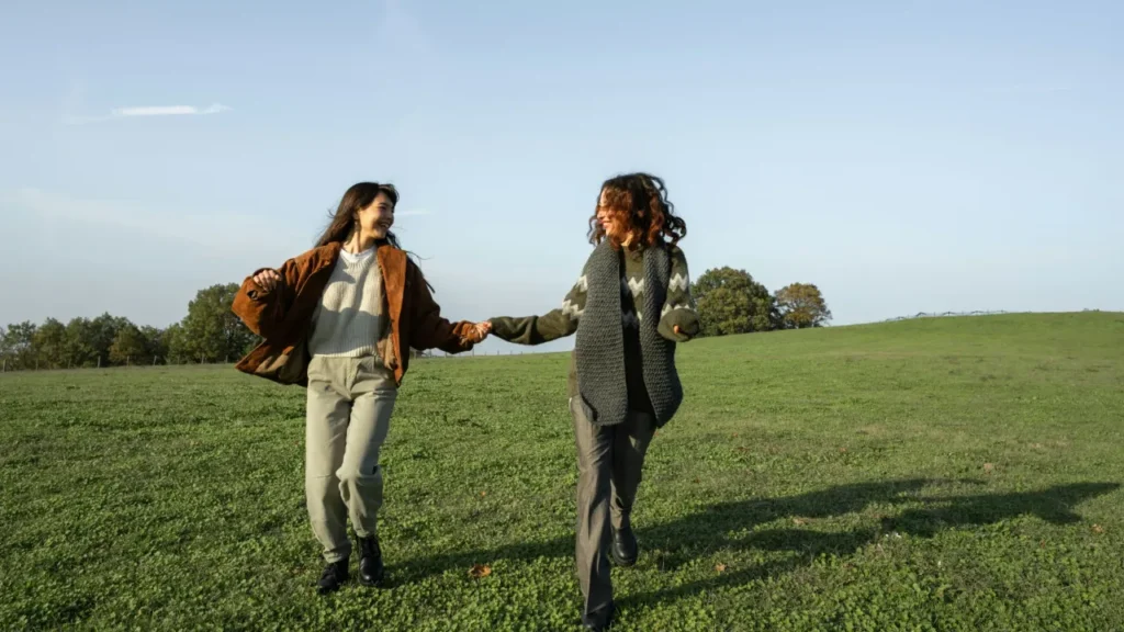 Two young women holding hands and laughing in a green field on a sunny day. A2 lesson plan friends.