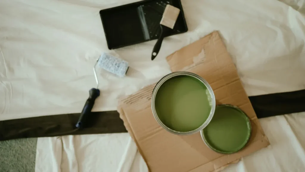 Two open cans of olive green paint with a roller and brush on a drop cloth, ready for a B1 lesson plan on home renovation.