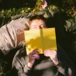 Person lying on grass reading a yellow book outdoors, surrounded by dandelions. B2 lesson plan books activity on why we should read.