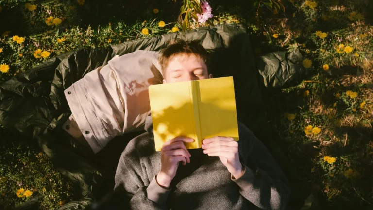 Person lying on grass reading a yellow book outdoors, surrounded by dandelions. B2 lesson plan books activity on why we should read.