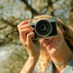 Woman holding a compact camera outdoors with blurred blossoms in background — b2 lesson photos on travel photography tips